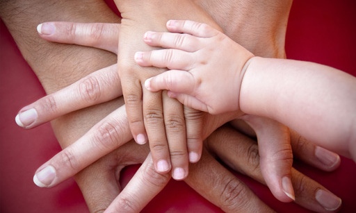 Four hands, one on top of the other. First two women’s hands, then a child’s hand, then a baby’s hand.