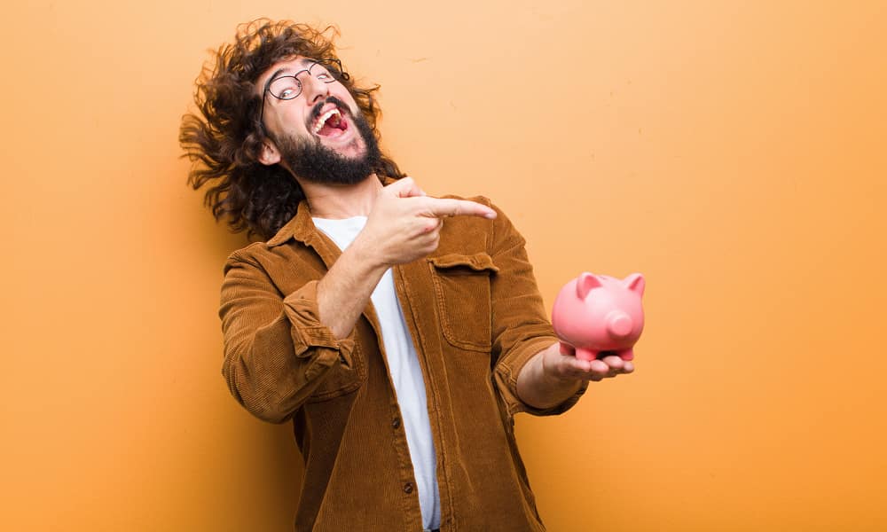 A white man with glasses, a beard, and long, curly hair, laughing and pointing at a piggy bank that’s in his hand.
