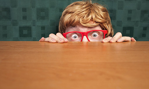 A man with ginger hair and red-framed glasses peeking from behind a wooden desk.