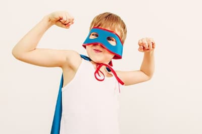 A young boy with a super hero mask and cape, flexing his muscles.