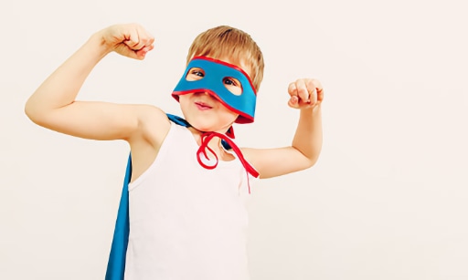 A young boy with a super hero mask and cape, flexing his muscles.
