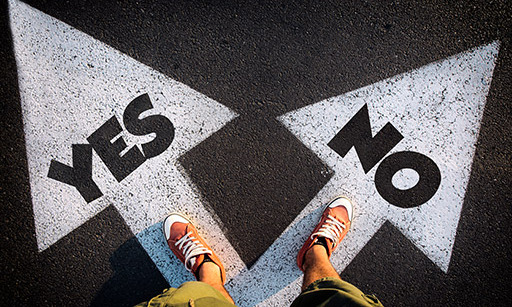 A man with shorts and trainers standing over a sign on the ground that consists of an arrow that forks into two, one with ‘Yes’ and the other with ‘No’.