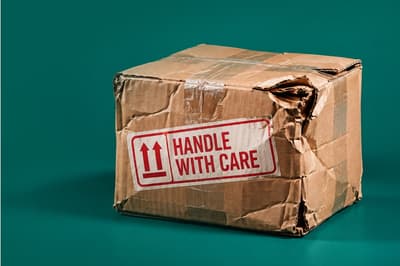 A battered cardboard delivery box with crushed corners and torn edges, featuring a prominent red and white ‘HANDLE WITH CARE’ label. The damaged parcel sits against a dark teal background.